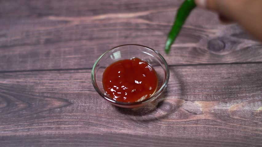 A Green chili dipped into a bowl of red sauce on a wooden table in a kitchen setup