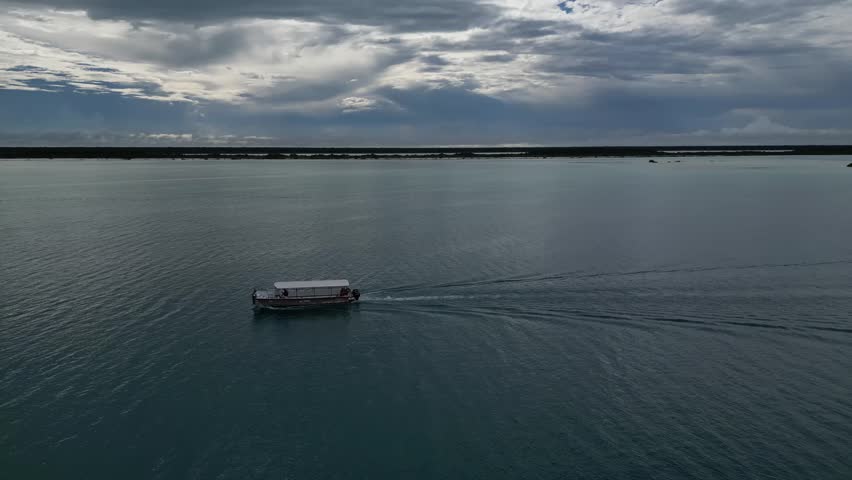An aerial view following a small boat moving across Bacalar Lagoon, leaving gentle ripples on calm blue water beneath a clear afternoon sky