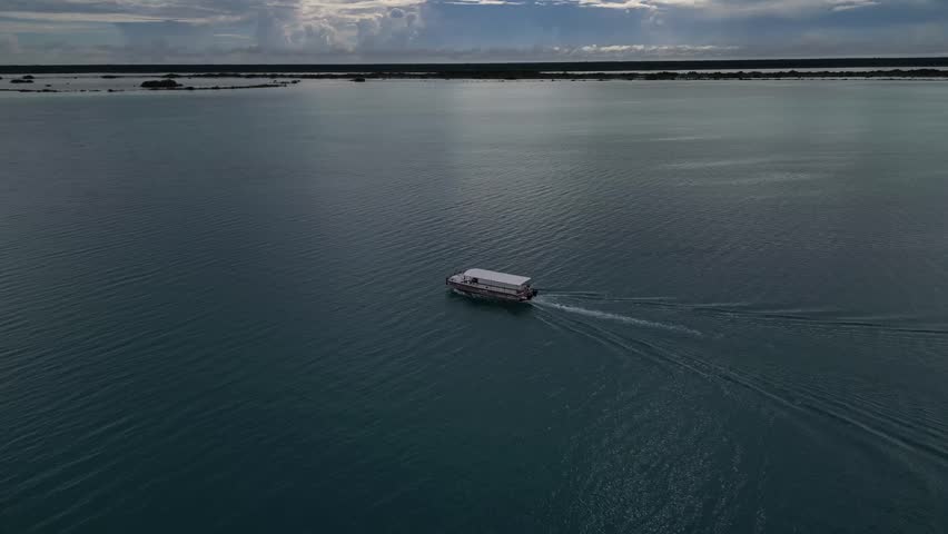 An aerial view tracking a small boat crossing Bacalar Lagoon, its soft wake spreading across calm blue water under a bright afternoon sky