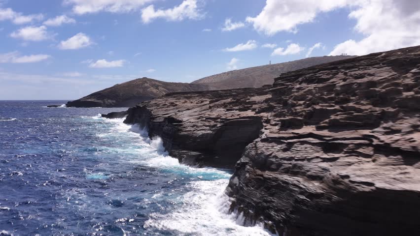 Cinematic drone footage flying over the ocean at Lanai Lookout in Hawaii on a sunny day. The aerial shot captures rugged rock formations as powerful