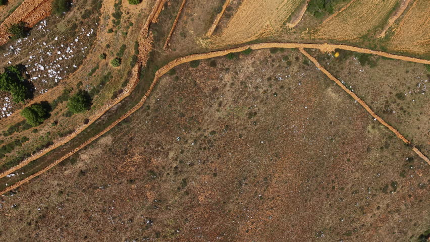 Overhead view of rural landscape showing harvested wheat fields alongside scrubland with scattered green trees, dry vegetation, and a dirt track running through the terrain