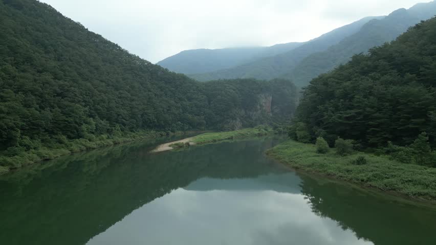 Serene landscape of a calm river reflecting surrounding mountains and dense forests in a remote South Korean valley. Perfect for themes of wilderness, tranquillity, and environmental conservation.