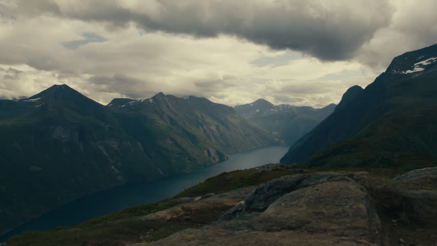 Rocky mountaintop ledge overlooks a long blue norwegian nature fjord framed by high dark slopes and a heavy cloud ceiling.