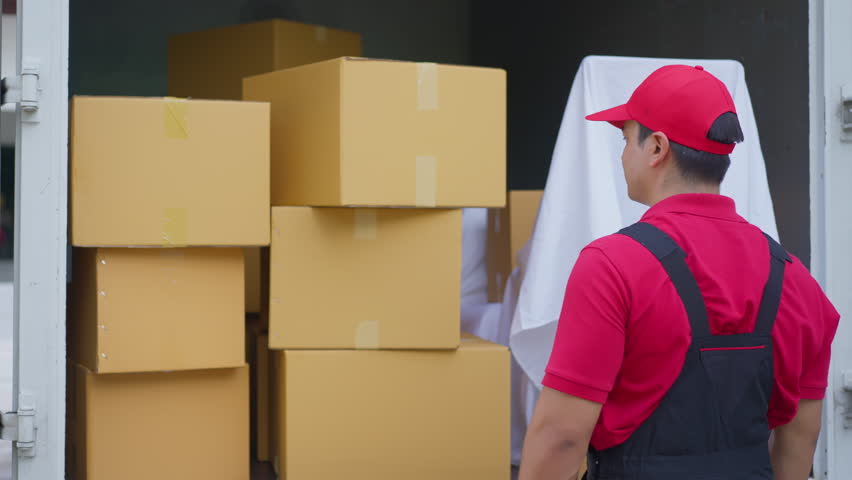 Delivery worker in red uniform with cap and smile moving cardboard box package in warehouse, courier service loading parcels into truck for relocation logistics