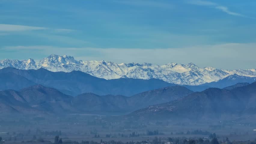 Telephoto panoramic pan across a jagged snow-capped Andean mountain range rising above layered blue foothills and a hazy valley floor under a vast open sky