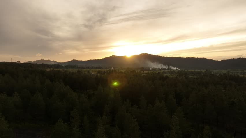 Warm sunrise light illuminates a misty mountain landscape with layered hills and soft fog drifting through the valleys. The scene shows a peaceful natural panorama with distant ridges, silhouetted trees, and a glowing morning sky.