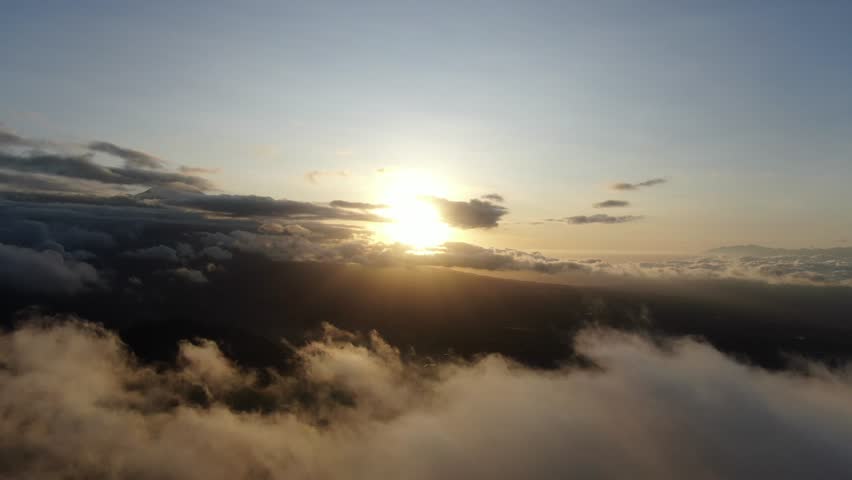 Warm sunrise light illuminates a misty mountain landscape with layered hills and soft fog drifting through the valleys. The scene shows a peaceful natural panorama with distant ridges, silhouetted trees, and a glowing morning sky.