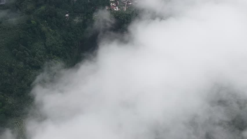 Aerial drone footage of a powerful sriti waterfall, lumajang, Indonesia cascading through a lush tropical forest, surrounded by rugged volcanic rocks and dense green vegetation. Mist rises from the rushing water, creating a dramatic and atmospheric natural landscape.