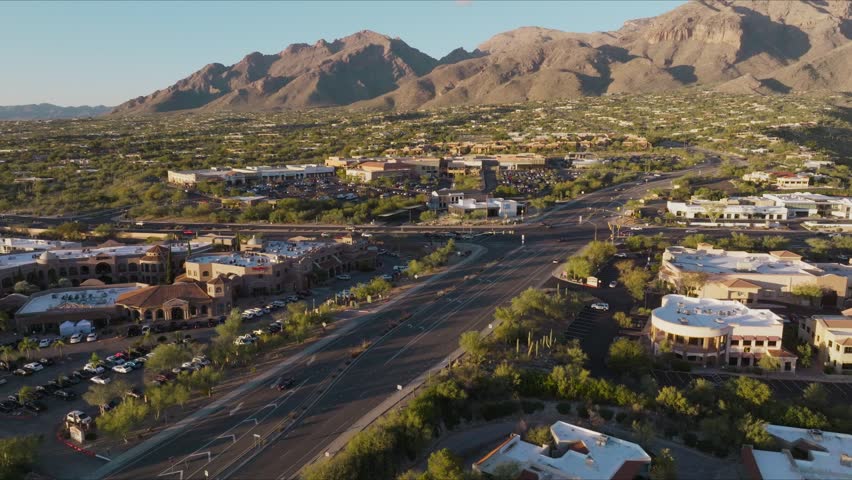 Drone footage captures traffic on highway intersecting with suburban retail area, mountains and sky at sunset.