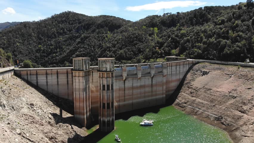 Dynamic aerial footage captures large dam structure with low water levels, revealing dry riverbed and surrounding vibrant green forest mountains under clear sunny sky, highlighting drought impact.