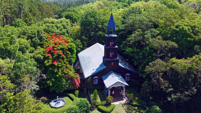 La Milagrosa Chapel rises with dark wood walls and blue steeple amid tropical forest in San Jose de las Matas Dominican Republic, flowering trees near quiet entrance. Aerial drone view.