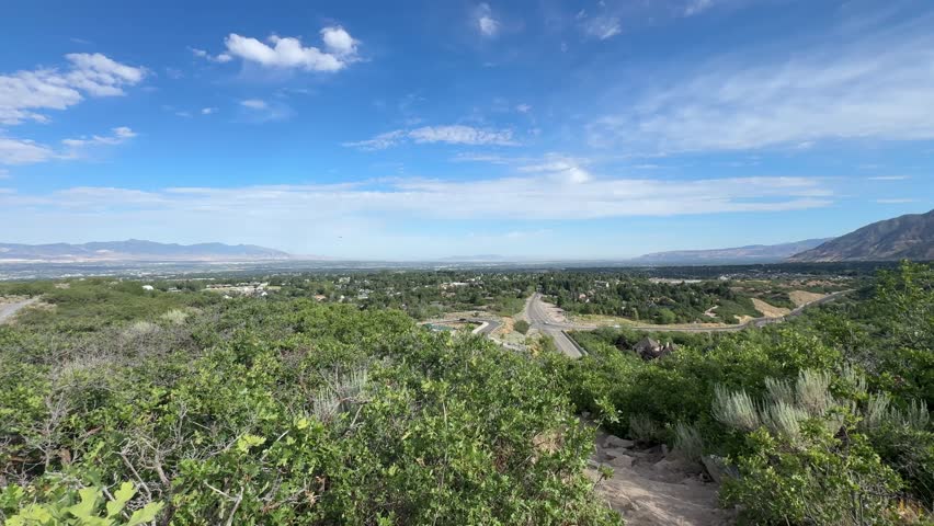 A wide elevated view over the Salt Lake Valley in Utah, looking across green foothills toward the urban sprawl and distant mountains. The scene fits the Salt Lake City area’s broad basin.