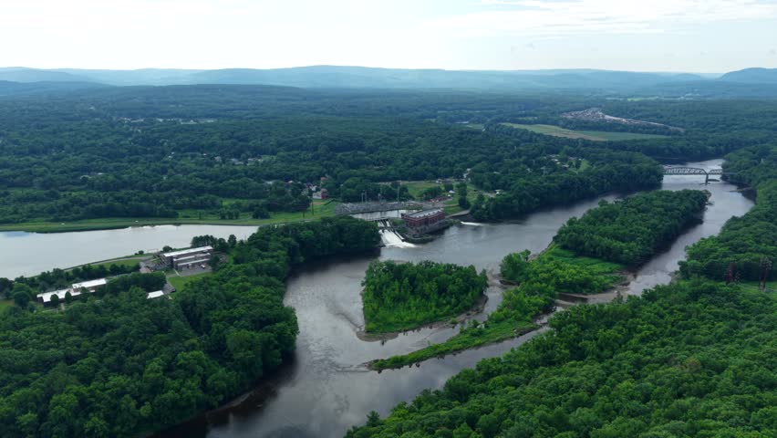 Connecticut River bends through forested landscape beside hydroelectric dam near Greenfield, Massachusetts, rolling hills stretching across scenic Pioneer Valley at dusk. Aerial drone wide shot