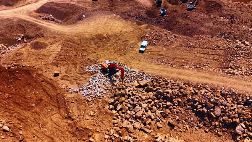 High angle aerial view of a stone pile and excavator in a quarry. An excavator stands near a large mound of loose rocks and a white van on dry ground.