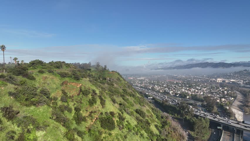 Dynamic aerial motion over green hillside revealing a bustling freeway with heavy traffic and dense urban landscape extending towards distant, misty mountains under blue sky with some clouds.