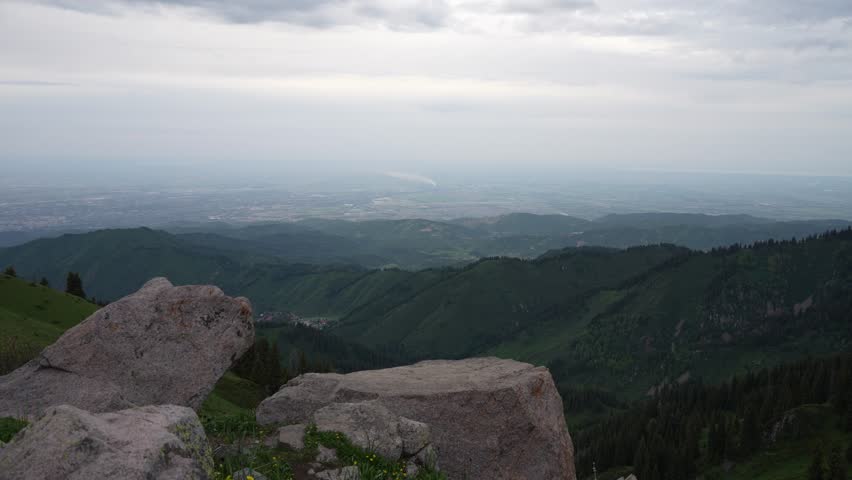 Scenic view of rocky foreground with large boulders overlooking lush green hills and valleys, under a cloudy sky, showcasing the natural beauty of the mountainous landscape. City of Almaty