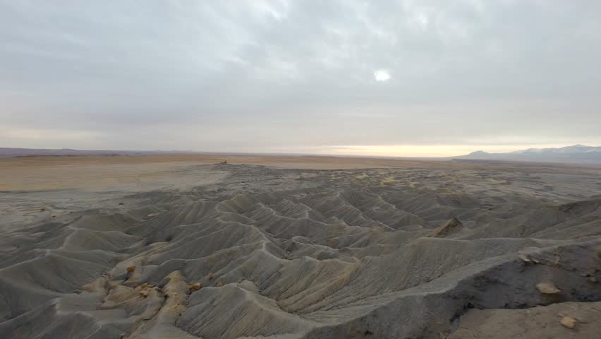 Cinematic aerial view flying low over textured grey desert hills in Utah badlands area under cloudy sunset sky capturing desolate barren terrain with cracked earth and sweeping motion.