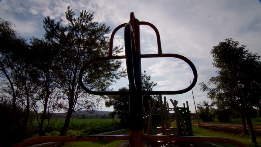 Backlit view of playground climbing bars in an outdoor park, with trees, grass and sunny sky creating a warm family recreation scene.