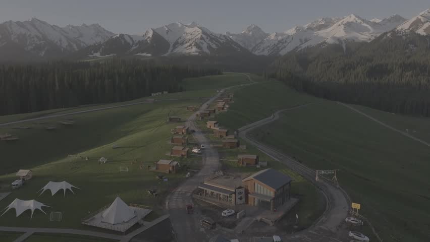 Drone view of luxury cabins surrounded by open meadows and snow capped mountains in Glen Etive Valley, showcasing a scenic alpine landscape with winding roads and evergreen forest