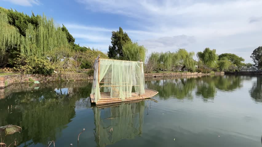 A high-quality 4K static shot capturing a traditional wooden raft adorned with flowing mint green curtains, resting peacefully on a calm pond within the historic Liyuan Garden in Wuxi, China. The scene features vibrant weeping willow trees and a clear blue sky reflected on the water surface. This serene and culturally rich footage evokes a sense of ancient Chinese elegance and tranquility, making it an ideal choice for travel documentaries, meditation backgrounds, or cultural heritage presentations.