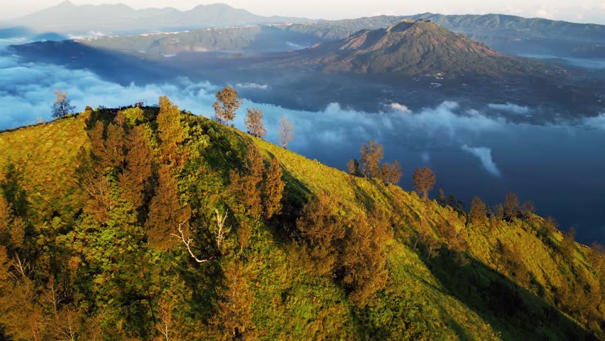 Drone flies over a grassy hillside with scattered trees, revealing a breathtaking panorama of a misty lake and distant volcanoes at sunrise.