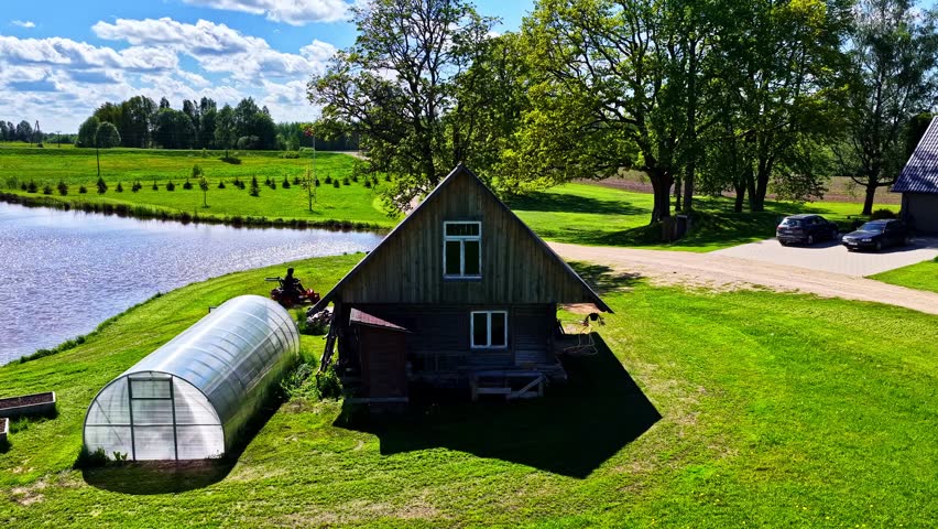 Aerial drone footage captured on summer day showing rustic timber cottage beside greenhouse, small pond, sprawling green lawns, tall leafy trees, and rural driveway with parked cars.