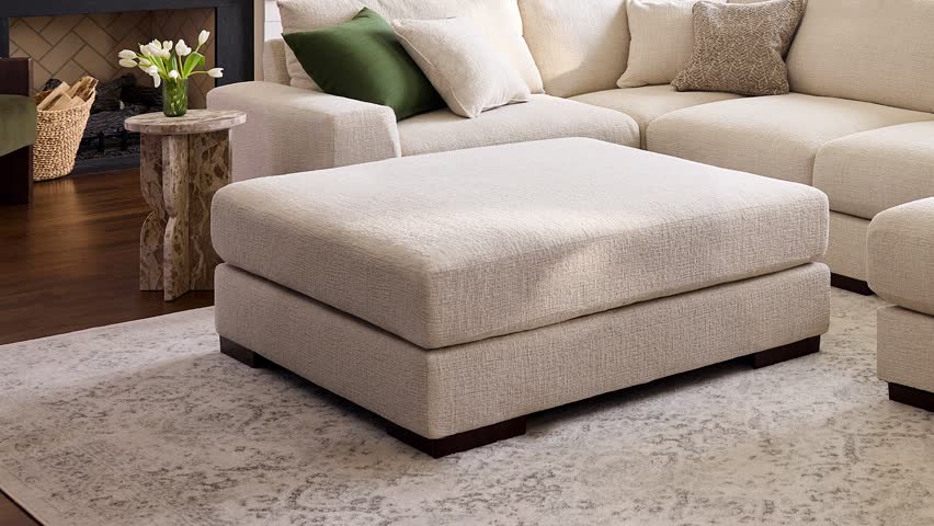 Slow zoom in on a large beige upholstered ottoman in a professionally staged farmhouse living room with warm natural light