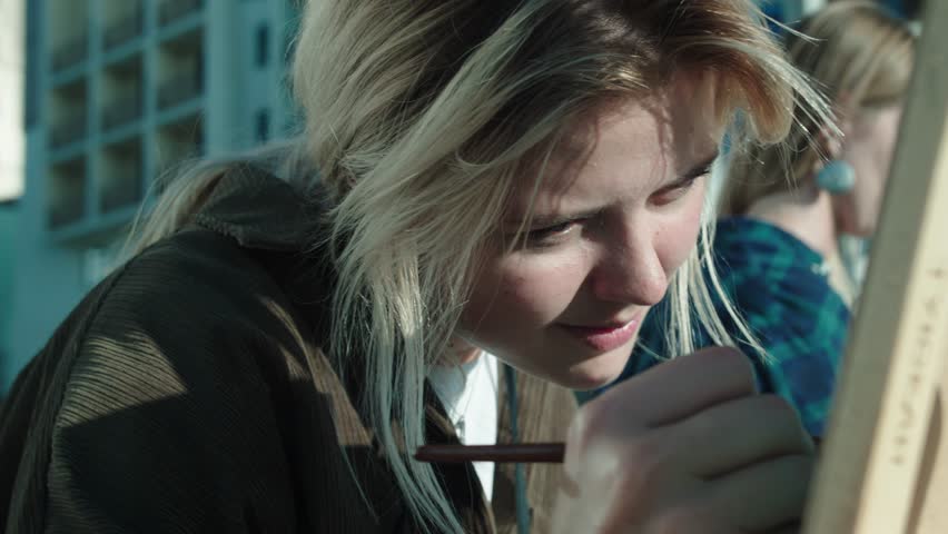 Blonde College of Arts student put her face close to easel. Carefully performs task of drawing with brush on easel with watercolors.
