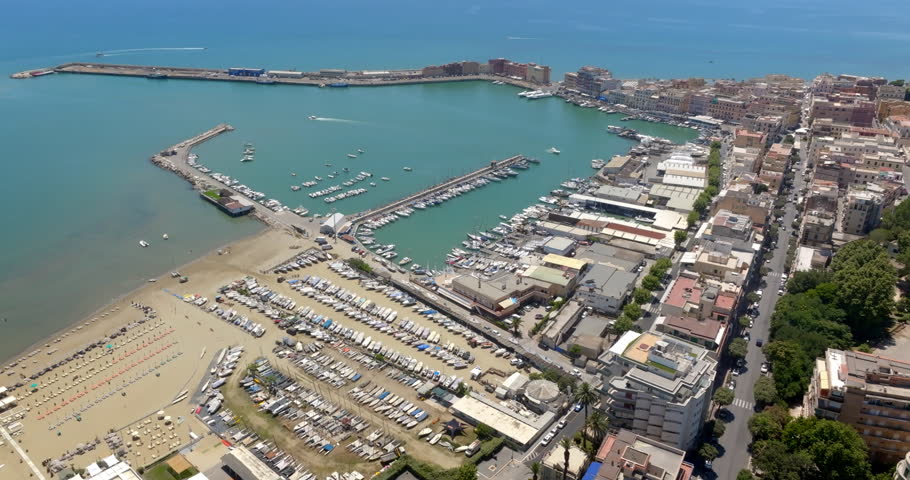 Aerial wide shot of the Anzio harbor and coastline, featuring vibrant sea waters, docked boats, sandy beaches, breakwaters, and urban coastal buildings in a summer day. It