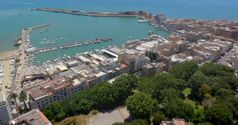 Aerial view of the harbor and coastal town of Anzio, located near Rome in Italy, featuring clear turquoise waters, breakwaters, docks with boats, and densely clustered buildings in a sunny summer day.