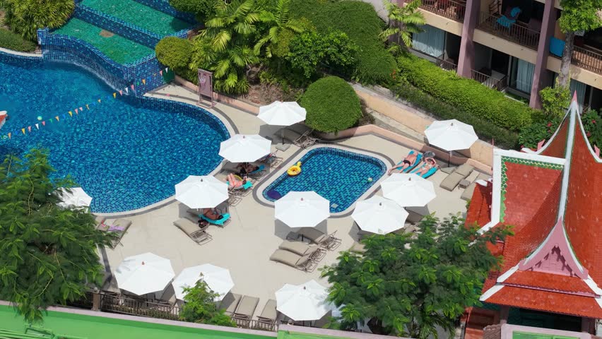 Aerial view of a vibrant resort pool area in Thailand, showcasing multiple swimming pools, lounge chairs, and umbrellas arranged around the water, with lush greenery in the background