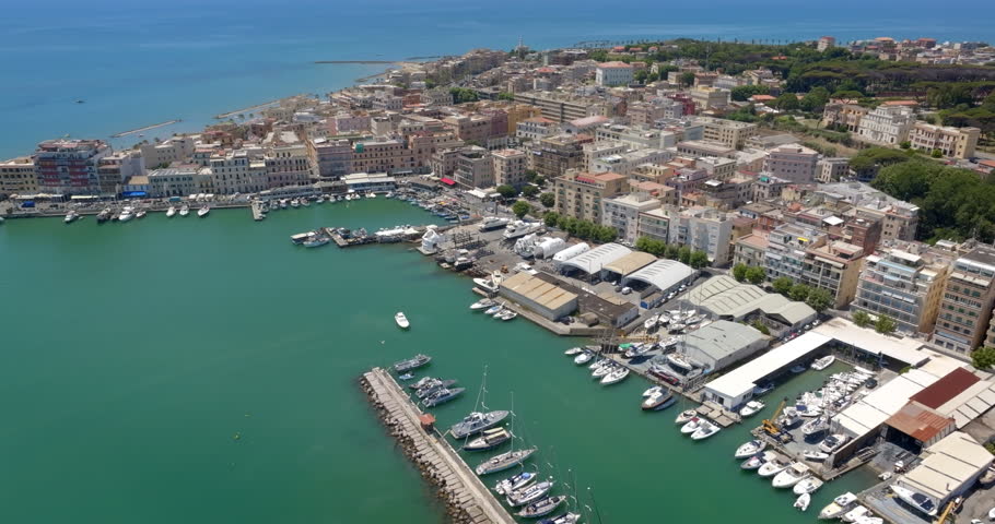 Aerial view of the coastal town of Anzio, near Rome in Italy, showing the bustling port with boats and yachts, turquoise sea waters, breakwaters, and colorful urban buildings along the coastline.