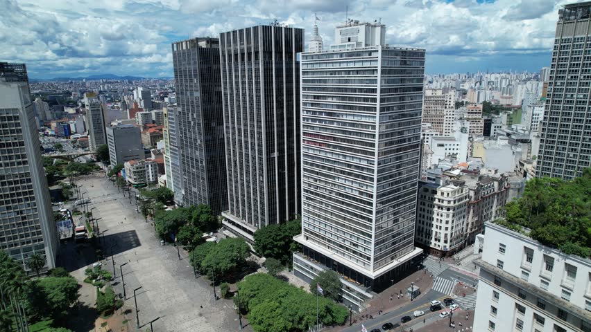 An aerial view shows the core of Sao Paulo with numerous buildings and streets. The city stretches wide under a clear sky with clouds. This view gives a sense of the city