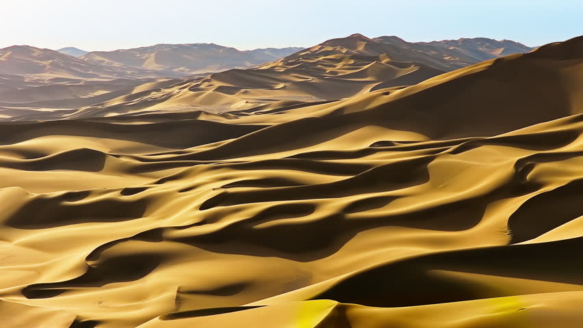 Aerial view of vast golden sand dunes stretching toward distant mountains under bright sunlight in Xinjiang.