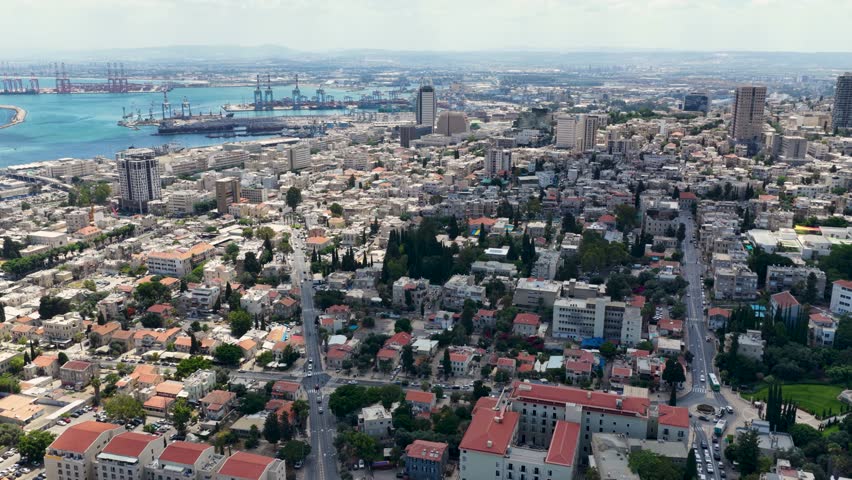 Cinematic drone footage showcasing the terraced Bahai gardens on Mount Carmel overlooking the dense urban sprawl and coastal neighborhoods of Haifa city on a clear summer day