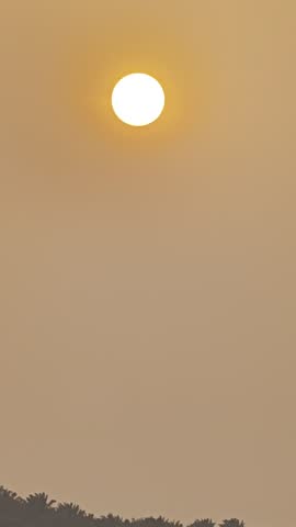 Bright glowing sun setting against a deep red orange sky over distant tree silhouettes, dramatic dusk landscape