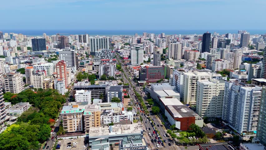 Cars driving on busy avenue surrounded by dense urban buildings and ocean in background in Santo Domingo, Dominican Republic. Aerial forward
