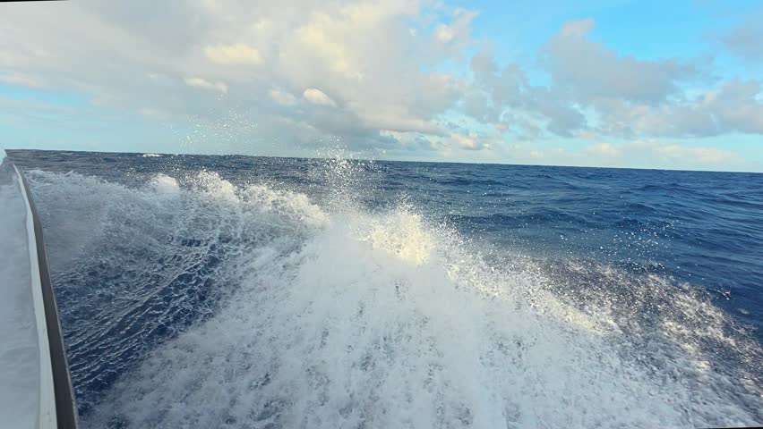 View from a boat moving fast across the deep blue ocean with water splashing and white foam against the hull under a cloudy sky