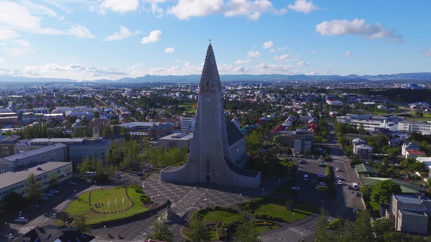 Aerial view of Hallgrimskirkja in Reykjavik, Iceland, gentle approach and pan show the stepped spire, plaza, Leif Erikson statue, city blocks, ports, and mountains.