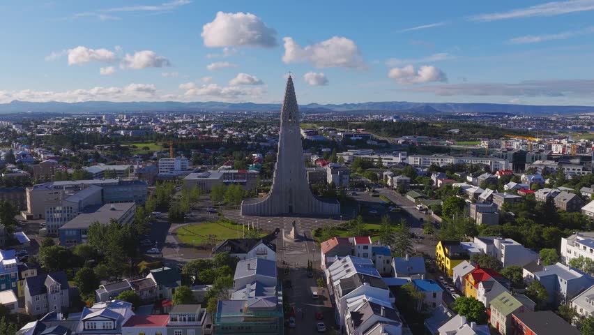 Aerial drone pan over Reykjavik, Iceland shows Hallgrimskirkja, painted houses, civic buildings, green plaza, harbor and mountains under sun and scattered clouds.