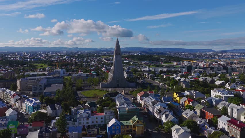 Aerial daytime view of Reykjavik, Iceland. Hallgrimskirkja rises over colorful houses as the camera glides forward and pans to the port, runways, and distant mountains.