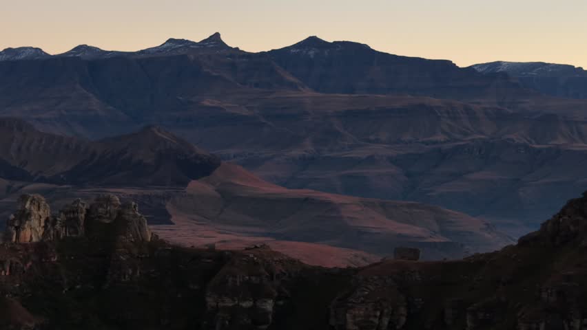 Scenic view of a mountain valley at dusk featuring multiple layered ridges and peaks silhouetted against a warm evening sky. Rocky cliffs frame the foreground with rolling hills extending into the distance.