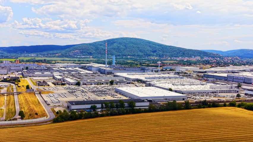 Factory with mountains behind. A large factory complex sits near a mountain. Trucks are parked in the lot under a clear sky. Fields surround the area.