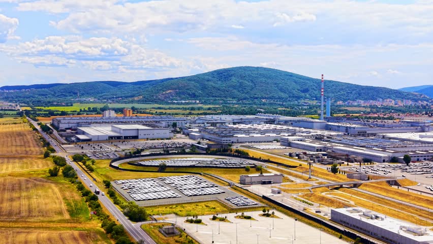 Factory by a big mountain. A large factory complex is set against a mountain in the background. Fields surround the area with some vehicles parked.