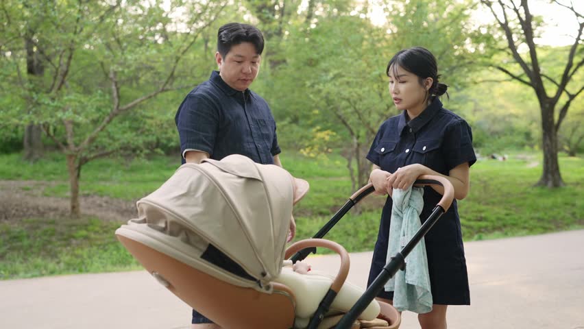 Couple walks through a park with a baby carriage. The man pushes the carriage gently. The woman holds a towel, watching nearby. They appear calm and content. Peaceful family outing