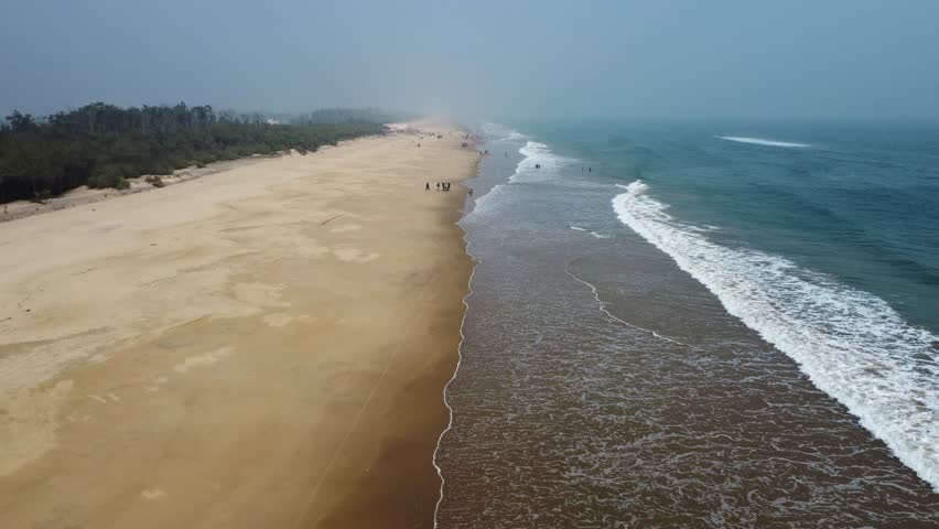 An aerial view of a long sandy beach with gentle waves and small groups of people walking along the shoreline