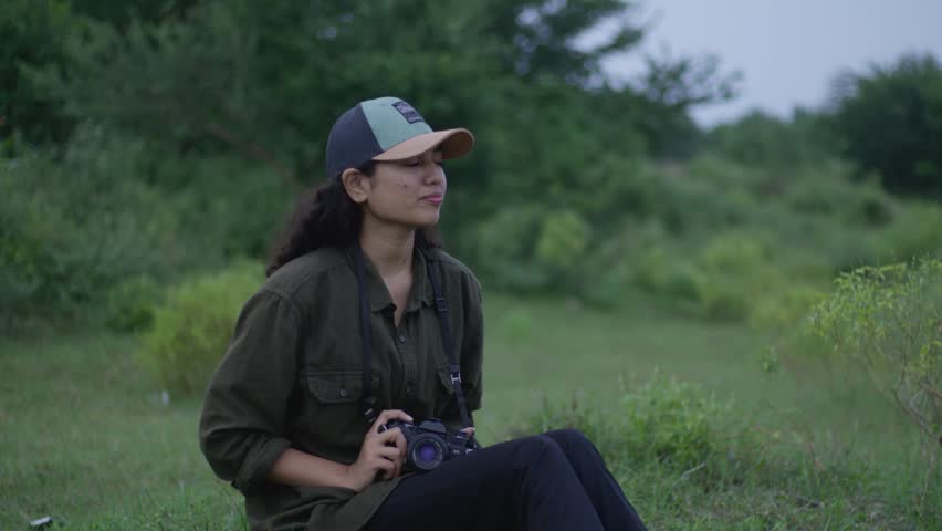 A seated portrait of a South Asian woman photographer holding a camera near the Kunwari River in Maharashtra, India, surrounded by greenery