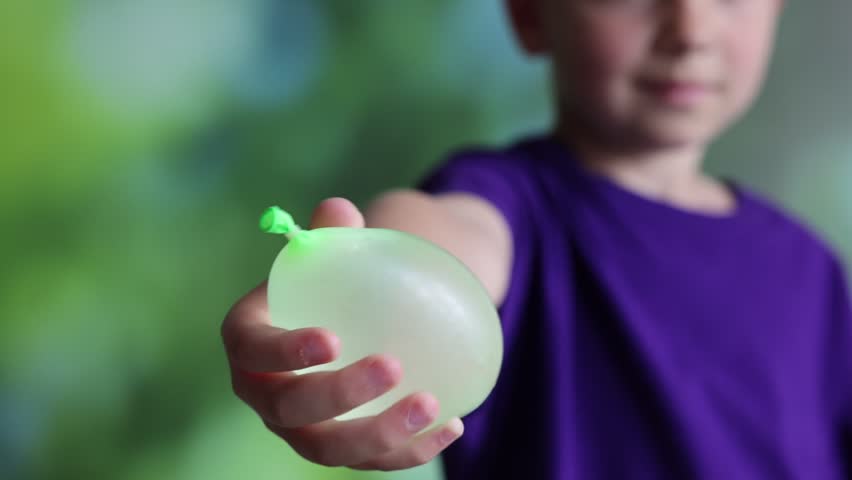Little boy holding green water balloon on blurred background, closeup. Camera moving up
