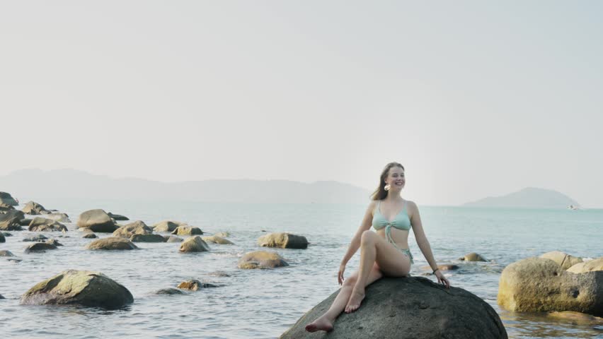 Female photo model in bikini is sitting on the stone on ocean beach and posing during session. Backstage of young adult pretty smiling woman taking part in shooting beauty pictures on the coastline