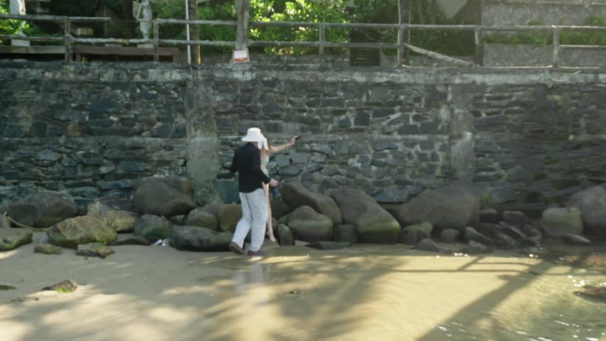 Backstage of bikini photo session on the beach with pretty Caucasian woman and male photographer. Slow motion of behind the scene process of taking pictures on sandy beach on stone wall background